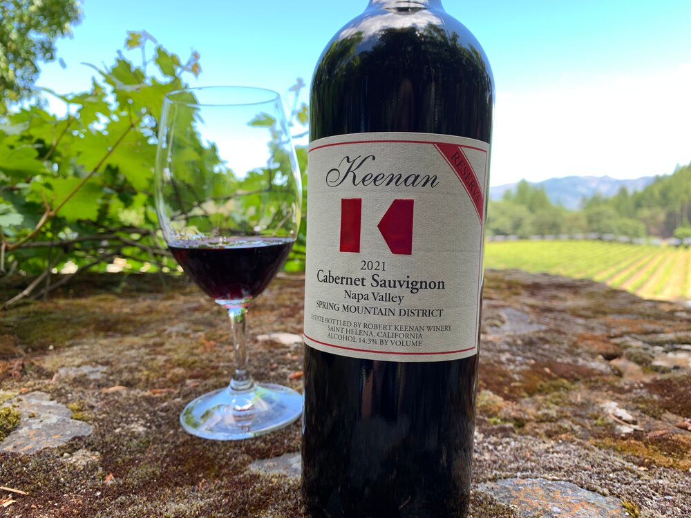 A bottle and poured glass of Keenan 2021 Cabernet Sauvignon Reserve, Spring Mountain District, on a rock above the estate vineyard