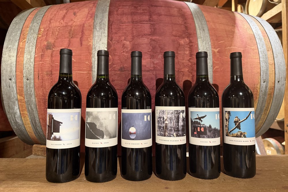 Six Keenan Bird Series bottles against a barrel — Raven 2020, Black Phoebe 2021, Northern Flicker 2022, Goose 2023 and Red-Tailed Hawk — each label carrying a photograph of its bird