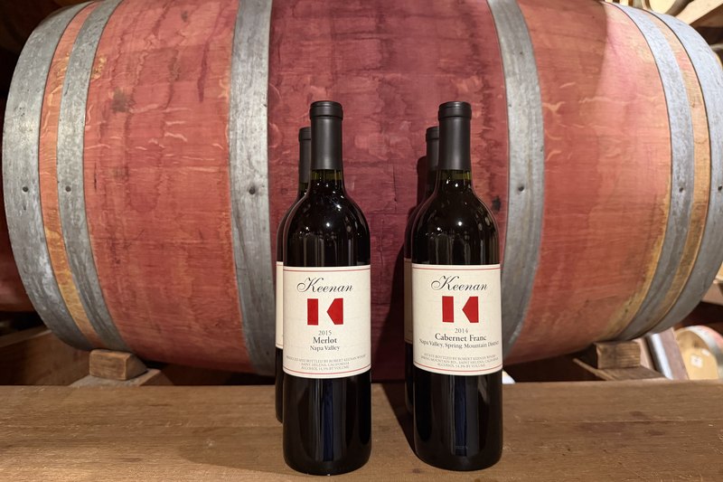 The Keenan Library Wine Club shipment against a barrel — two bottles each of the 2015 Merlot and the 2014 Cabernet Franc