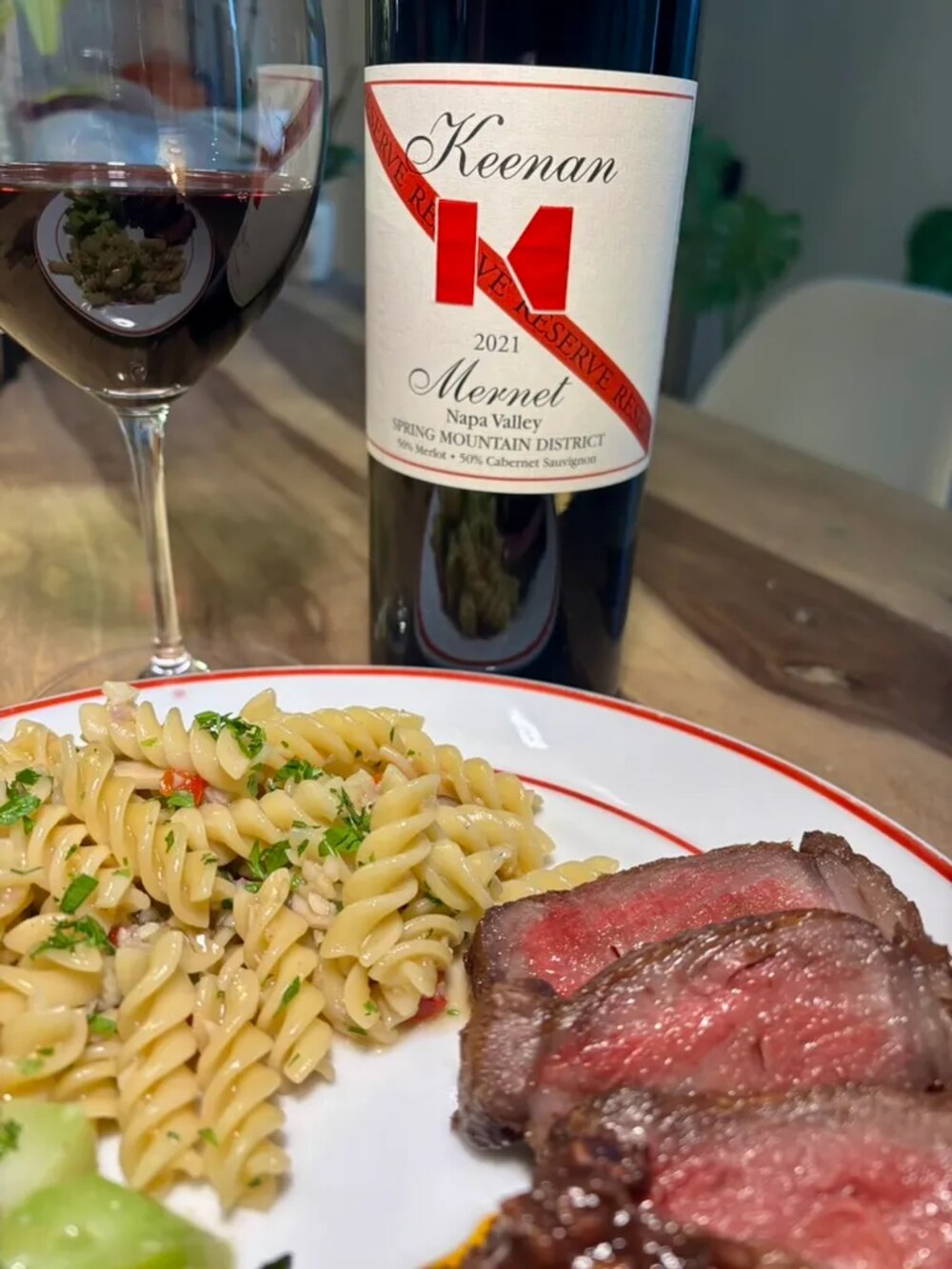 A sliced Vermont Wagyu New York strip plated with pasta, beside a glass and bottle of Keenan 2021 Mernet Reserve