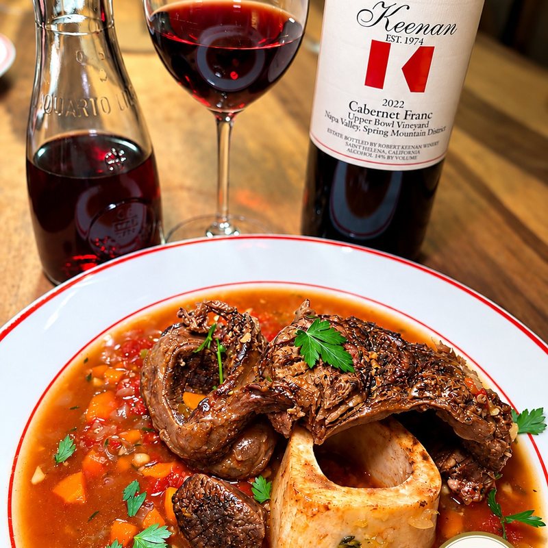 Braised Vermont Wagyu osso buco with its marrow bone in a tomato and vegetable sauce, beside a glass, a carafe and a bottle of Keenan 2022 Cabernet Franc, Upper Bowl Vineyard