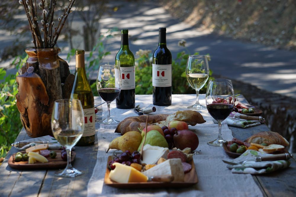 Keenan bottles and glasses on an outdoor table with a board of cheese, charcuterie, bread and fruit