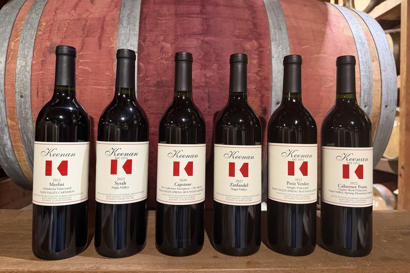The Keenan Reds Only Club shipment against a barrel — Merlot, Syrah, Cabernet Sauvignon, Zinfandel, Petit Verdot and Cabernet Franc