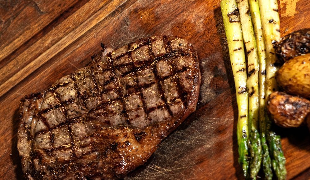 A cooked rib eye steak resting on a wooden board with charred asparagus and roast potatoes