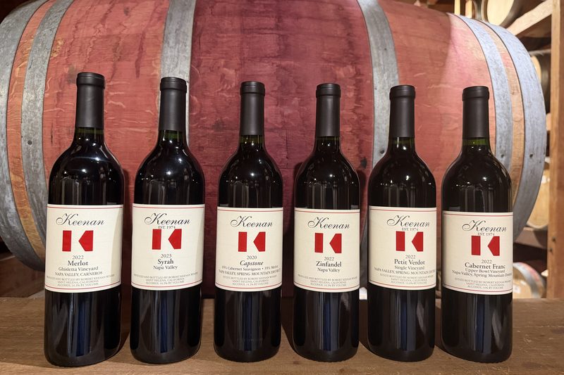 The Keenan Wine Club shipment against a barrel in the cellar — six red bottles: Merlot, Syrah, Capstone, Zinfandel, Petit Verdot and Cabernet Franc
